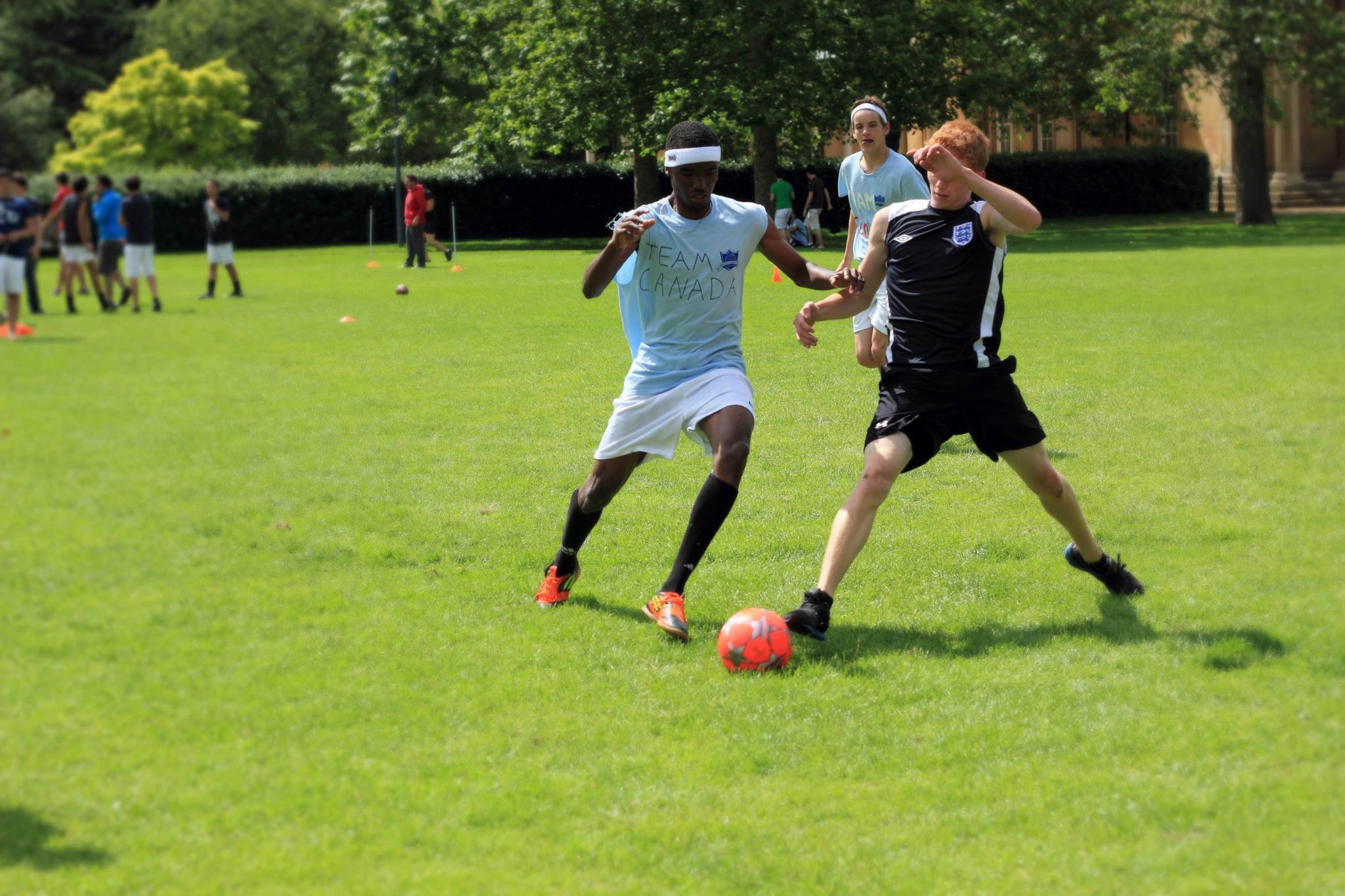 Two students play football on a green field. One student has red hair and is wearing their own sports kit, and the other student is wearing a customised 'Reach Cambridge' T-shirt. They are both running for the ball.