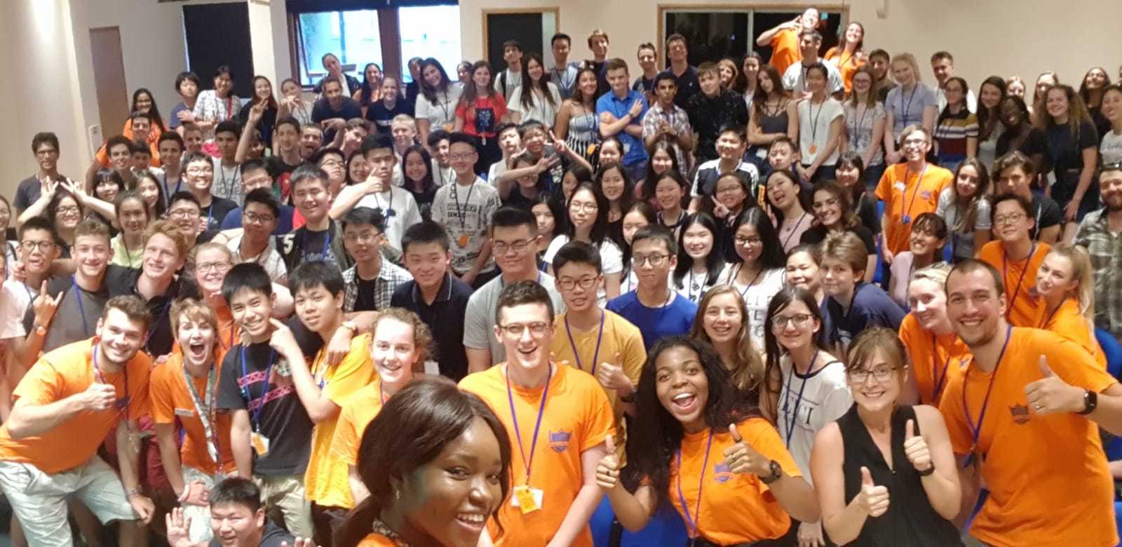 A wide-angle selfie of all of the students from a summer program. The Reach staff are at the front in orange T-shirts, looking happy and excited. The students are all standing behind, smiling.