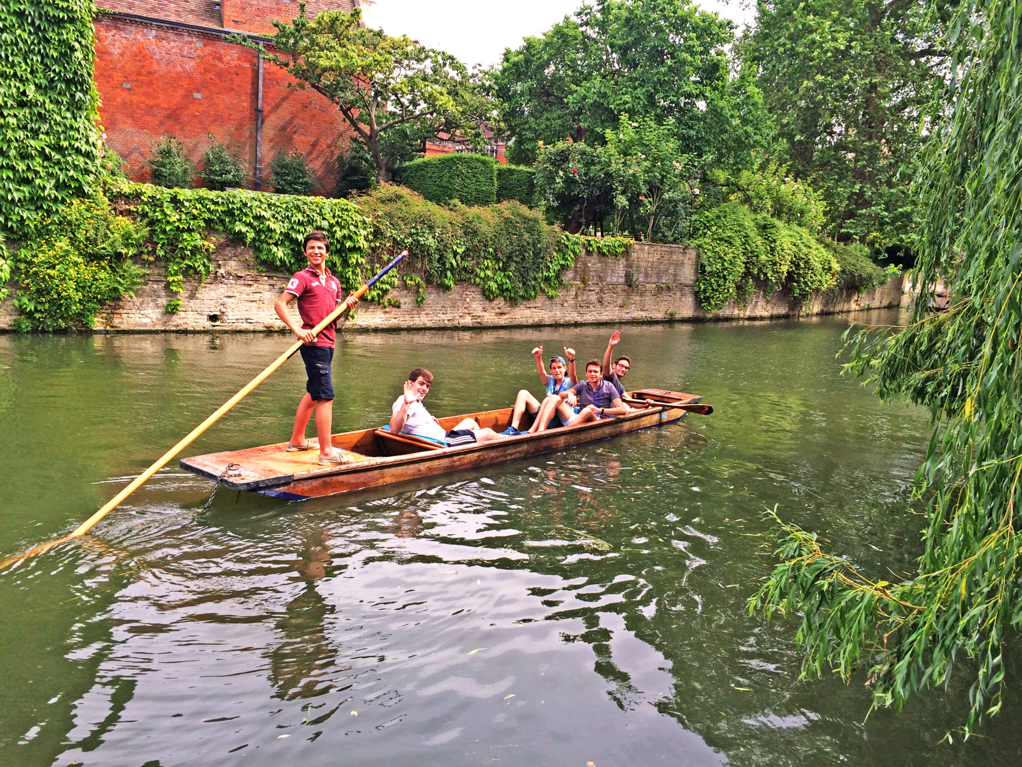 A group of students punting on the River Cam. One student stands at the back of the punt holding a pole. The others are sat in the boat, waving at the camera.