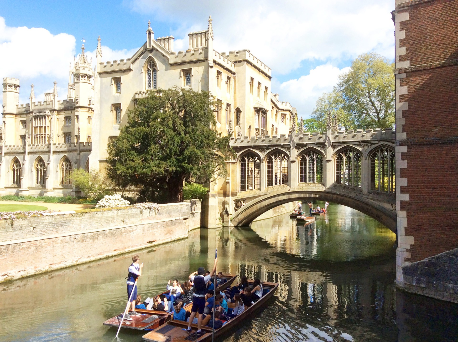 Students punting on the River Cam under the historic Bridge of Sighs at St John's College, Cambridge, on a sunny day.