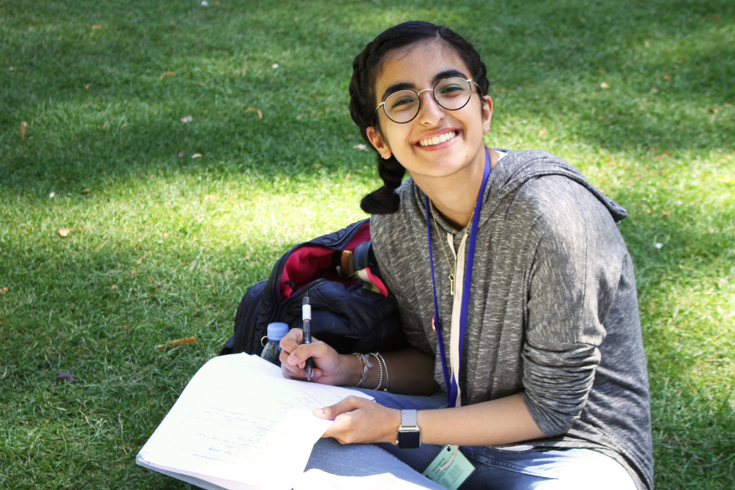 A smiling student sitting on the grass, writing in a notebook during a sunny day at Reach Cambridge.