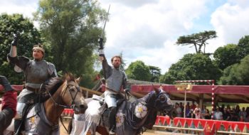 Two knights in armor riding horses and raising their swords during a live historical re-enactment at Warwick Castle, with spectators watching in the background.