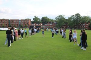 A large group of students standing in a line on a green field, engaging in a social activity on a sunny day, with residential buildings in the background.